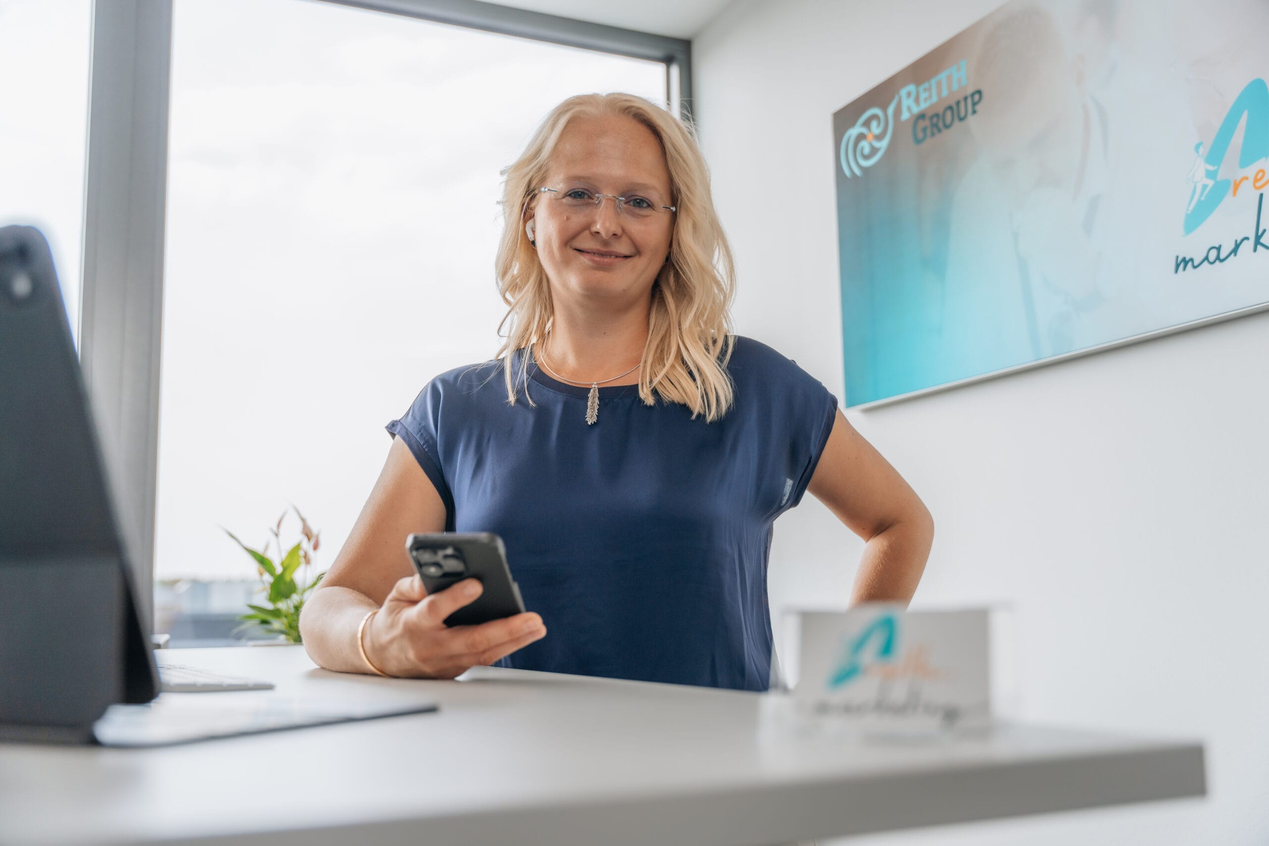 Justine Reith steht am Schreibtisch mit Blick in die Kamera, während sie ihr iPhone hält und AirPods trägt.